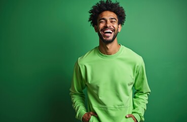 Handsome Black man laughs heartily wearing bright lime green sweatshirt against solid green studio background. Curly afro hair, beard. Man stands with hands in pockets radiating joy, confidence.