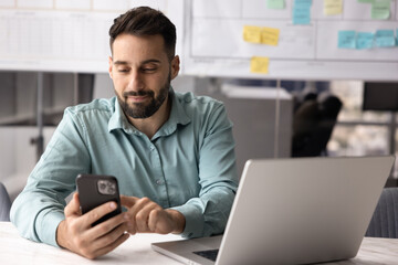 Young Latin businessman sit at desk with open laptop using his modern smartphone during break,...