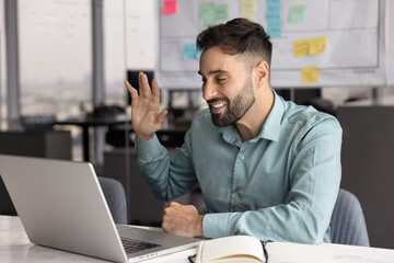 Positive professional businessman sits at desk looks at laptop screen greeting client start online...