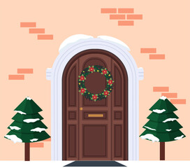 Fragment of the house facade with a door and a Christmas wreath. Christmas trees at the entrance. Brick facade. Festive atmosphere and anticipation of Christmas and New Year. Winter festive decoration