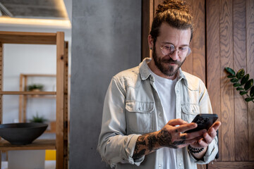 Young man with tattoos and glasses using smartphone in modern apartment