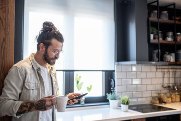 Freelancer enjoying morning coffee and checking smartphone in modern kitchen