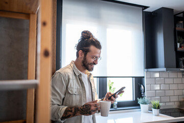 Freelancer enjoying morning coffee and checking messages on smartphone in kitchen