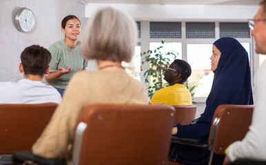 Emotional young female tutor leading educational class for multiethnic adult group sitting in...