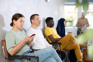 Young woman in casual clothes waiting for her turn while sitting on chair with phones in her hand