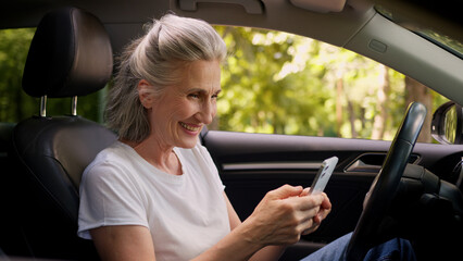 Cheerful Caucasian mature woman elderly senior female retired pensioner car automobile driver inside auto using mobile phone playing game clench fist celebrating win victory betting success offer deal