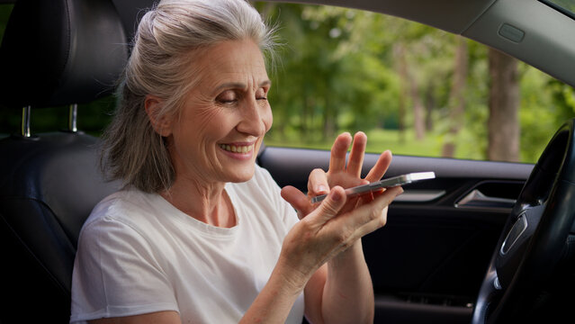Caucasian grandmother woman female senior driver mature car rental service client user holding mobile phone using artificial intelligence assistant speech app record voice message inside automobile