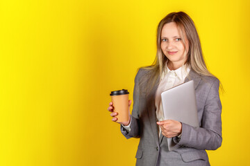 Smiling happy young businesswoman carrying laptop computer and cup of coffee to go against yellow...