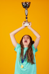 Happy young blonde girl with a gold medal holding winner cup or trophy on yellow background. She is...