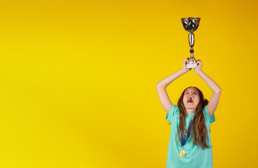 Happy young blonde girl with a gold medal holding winner cup or trophy on yellow background. She is...