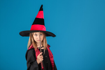 Halloween Theme. Young witch girl in black mantle, hat and magic wand against blue background.