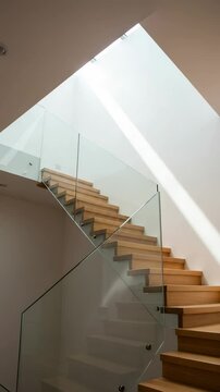 Minimalist wooden staircase with a frameless glass banister in a modern house. Architectural detail with natural light from a skylight.