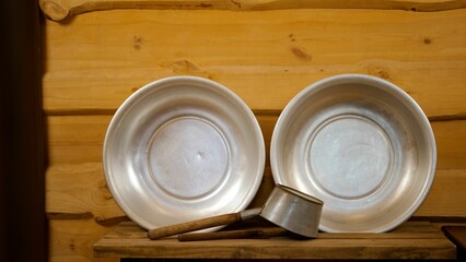 Metal washbasins and a ladle sit on a wooden shelf against a wooden wall, creating a rustic and traditional bathing scene