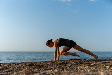 Fit woman wearing black activewear performing mountain climbers exercise on a pebbled beach at sunrise, focusing on strength, cardio, and endurance as part of a healthy outdoor fitness routine