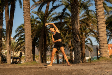 Slim athletic woman in a black outfit performing a side stretch exercise barefoot under tropical...