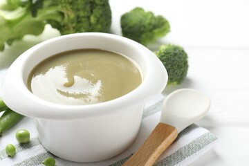 Baby food. Tasty puree in bowl, spoon, peas and broccoli on white wooden table, closeup