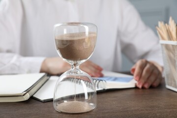 Deadline. Woman with notebooks working at wooden table in office, focus on hourglass