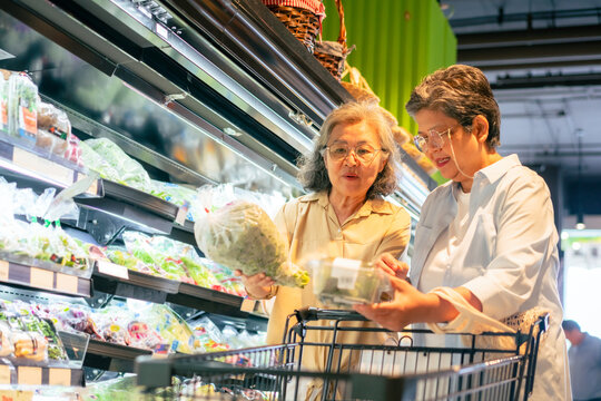 Happy Asian senior woman shopping buying food and grocery at supermarket. Elderly women friends enjoy healthy lifestyle choosing and buying organic fresh food, vegetable and fruit at shopping mall.