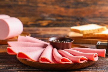 Plate with slices of tasty boiled sausage, bread and peppercorns on brown wooden background