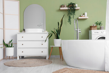 Bathtub, houseplants and chest of drawers near green wall in interior of bathroom