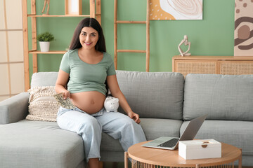 Young pregnant woman with piggy bank and money sitting on sofa at home. Maternal Benefit concept