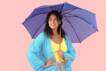 Happy young African-American woman in raincoat with umbrella on pink background