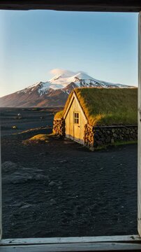 View from a window of a traditional Icelandic turf house. Scenic landscape with a snow-capped volcano and black sand during golden hour.