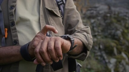 Medium midsection shot of hands of anonymous male hiker in khaki shirt, with backpack and trekking poles checking compass on smart watch for direction bearings while walking in mountains - Powered by Adobe
