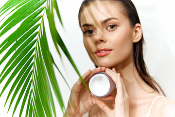 Young woman with natural beauty holding skincare cream, framed by green palm leaves, showcasing...