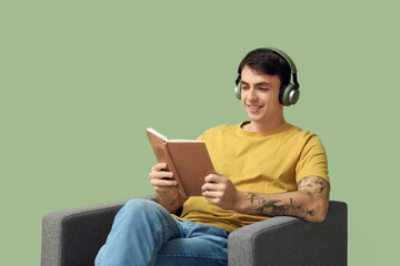 Handsome young man in headphones reading book on armchair against green background
