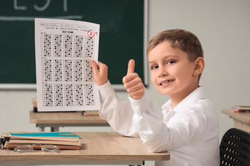 Cute schoolboy with results of test showing thumb-up at desk in classroom