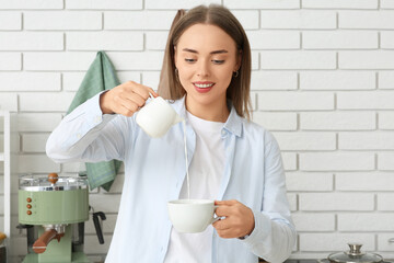 Beautiful young woman pouring fresh milk from jug into cup of hot coffee in kitchen