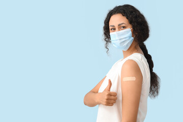 Young African-American woman in mask with medical patch showing thumb-up after vaccination on blue...
