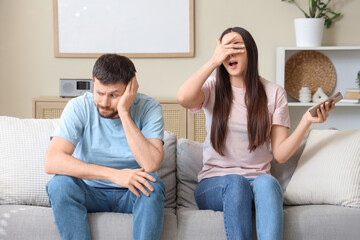Shocked young woman with mobile phone and her husband quarreling at home