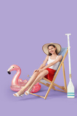 Young woman with summer hat sitting in deck chair on lilac background