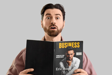 Shocked young man reading business magazine on light background