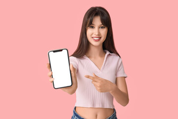 Smiling young woman pointing at blank mobile phone on pink background