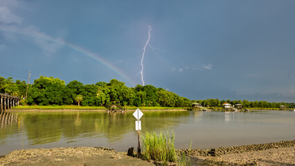 Bright lightning strikes over a calm river with rainbow and lush green trees in the background...