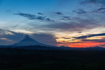 Mount Agung volcano in Bali, Indonesia, is silhouetted against a vibrant sunset sky. The landscape...