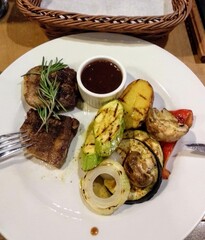 Grilled meat platter with vegetables and sauce served on a white plate in a restaurant setting