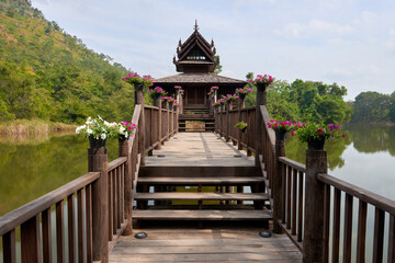 Obraz premium Stunning Wooden Bridge Leading to a Traditional Structure by a Serene Lake Surrounded by Lush Greenery