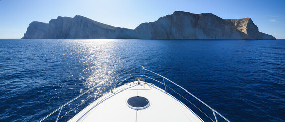 The bow of a sleek yacht cuts through calm ocean waters under a bright sky. The view from the front of the boat captures a feeling of freedom, travel, and endless adventure at sea.