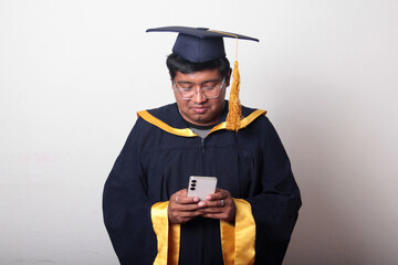 Young man dressed in cap and gown to celebrate his graduation uses his cell phone to take selfies,...