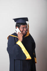 Young man dressed in cap and gown to celebrate his graduation uses his cell phone to take selfies,...