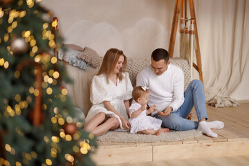 Young Caucasian parents in white clothes sit with baby girl on rug in light wooden room, Christmas tree in foreground. Front view. Concept: family, childhood, holidays, home comfort.