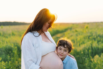 A pregnant woman stands in a lush green field, gently holding her baby bump. Her son leans against her, both sharing a tender moment amidst tall grass and a warm sunset.