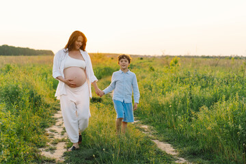 A pregnant woman stands in a lush green field, gently holding her baby bump. Her son leans against her, both sharing a tender moment amidst tall grass and a warm sunset.