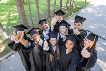 Students of different nationalities in graduation gowns give thumbs up. 