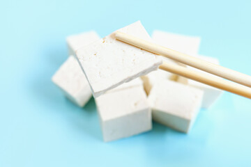 Piece of white tofu in wooden chopsticks on blue background close-up.