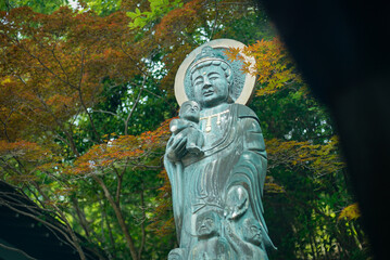 Fototapeta premium A peaceful bronze statue of Kannon, the Buddhist goddess of mercy, holding a child at Myoen-ji Temple in Japan. Surrounded by colorful trees in a tranquil garden setting, symbolizing compassion and sp